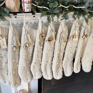 Op de afbeelding: Een rij cr&egrave;mekleurige gebreide kerstsokken met een verhoogde pompontextuur. Elke sok is versierd met een lichtblauw lint, een houten naamplaatje en twee kleine goudkleurige metalen belletjes die aan de bovenste lus hangen.