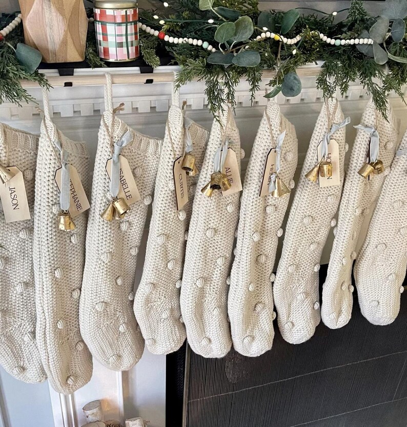 Op de afbeelding: Een rij cr&egrave;mekleurige gebreide kerstsokken met een verhoogde pompontextuur. Elke sok is versierd met een lichtblauw lint, een houten naamplaatje en twee kleine goudkleurige metalen belletjes die aan de bovenste lus hangen.