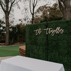 May include: A green artificial hedge backdrop with a wooden sign that reads "the trujillos" in cursive script. A white table sits in front of the backdrop.