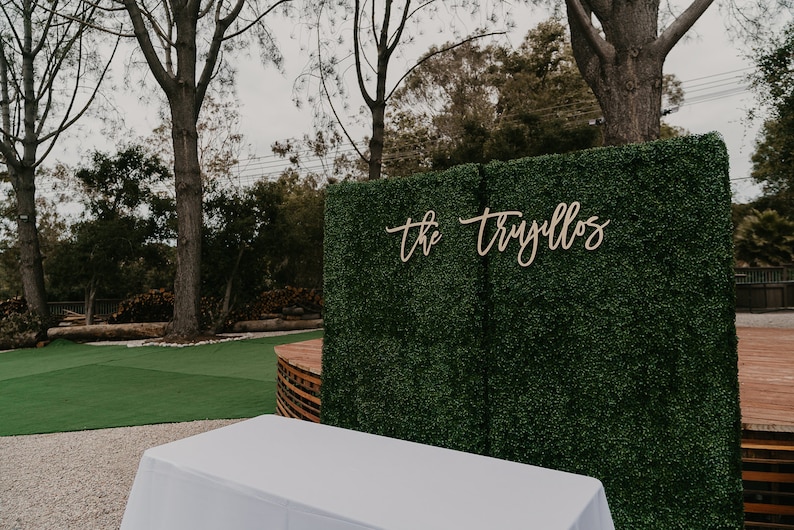 May include: A green artificial hedge backdrop with a wooden sign that reads "the trujillos" in cursive script. A white table sits in front of the backdrop.