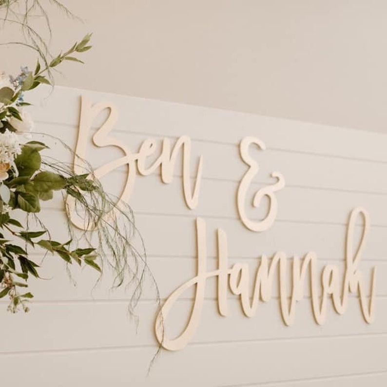 May include: Wooden sign with the names "Ben & Hannah" written in cursive script. The sign is made of light wood and is perfect for a wedding or engagement party.