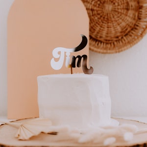 May include: A white cake with a silver 'Tim' cake topper on top. The cake is on a wooden slice with a brown background.