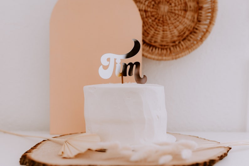May include: A white cake with a silver 'Tim' cake topper on top. The cake is on a wooden slice with a brown background.