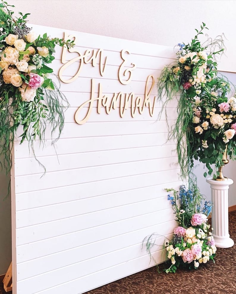 May include: White wooden backdrop with a sign that reads "Ben & Hannah" in cursive script. The backdrop is decorated with greenery and flowers.