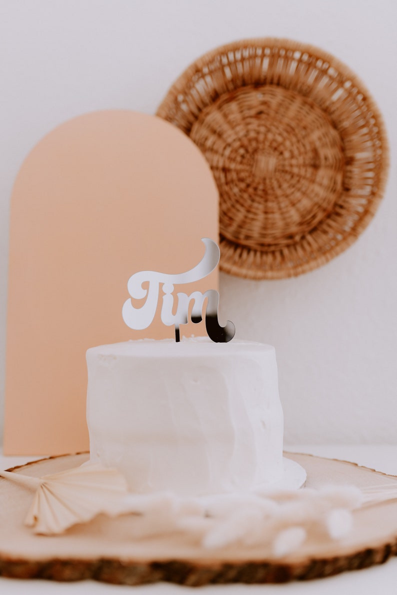 May include: A white cake with a silver 'Tim' topper on a wooden cake stand. The cake is in front of a peach-colored arch and a woven basket.