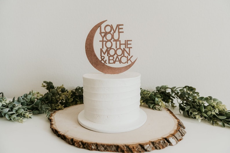May include: A white cake with a wooden cake topper that says "Love you to the moon & back" in a crescent moon shape.