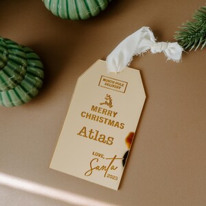 May include: Gold Christmas gift tag with the text "Merry Christmas Atlas Love, Santa 2023" and a small reindeer graphic. The tag has a white ribbon attached.