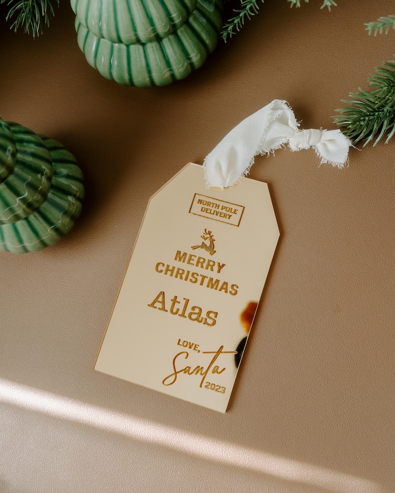 May include: Gold Christmas gift tag with the text "Merry Christmas Atlas Love, Santa 2023" and a small reindeer graphic. The tag has a white ribbon attached.