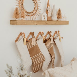 Op de afbeelding: Vijf kerstsokken in cr&egrave;me- en bruintinten hangen aan een houten harmonica-rek. De sokken hebben gebreide texturen en horizontale strepen, elk versierd met een rechthoekig houten naamplaatje. Het rek is bevestigd aan een witte muur.