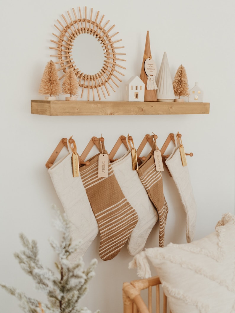 Op de afbeelding: Vijf kerstsokken in cr&egrave;me- en bruintinten hangen aan een houten harmonica-rek. De sokken hebben gebreide texturen en horizontale strepen, elk versierd met een rechthoekig houten naamplaatje. Het rek is bevestigd aan een witte muur.