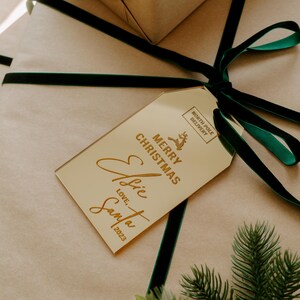 May include: A gold gift tag with the text "Merry Christmas" and "Ellie" written in cursive. The tag also includes the text "Love: Santa 2023" and a small stamp that says "North Pole Delivery".