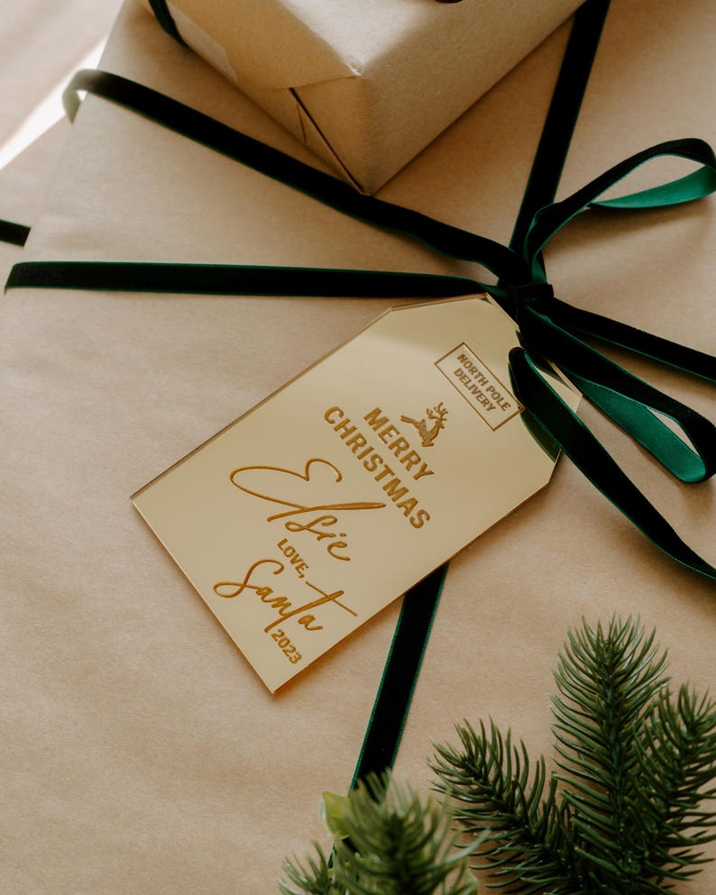 May include: A gold gift tag with the text "Merry Christmas" and "Ellie" written in cursive. The tag also includes the text "Love: Santa 2023" and a small stamp that says "North Pole Delivery".