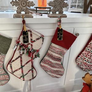 Op de afbeelding: Twee kerstsokken hangend aan een wit oppervlak. De linker sok is cr&egrave;mekleurig met een rood en groen ruitpatroon en franjes. De rechter sok heeft een effen rode boord en een cr&egrave;mekleurig lichaam met rode en groene geweven geometrische strepen.