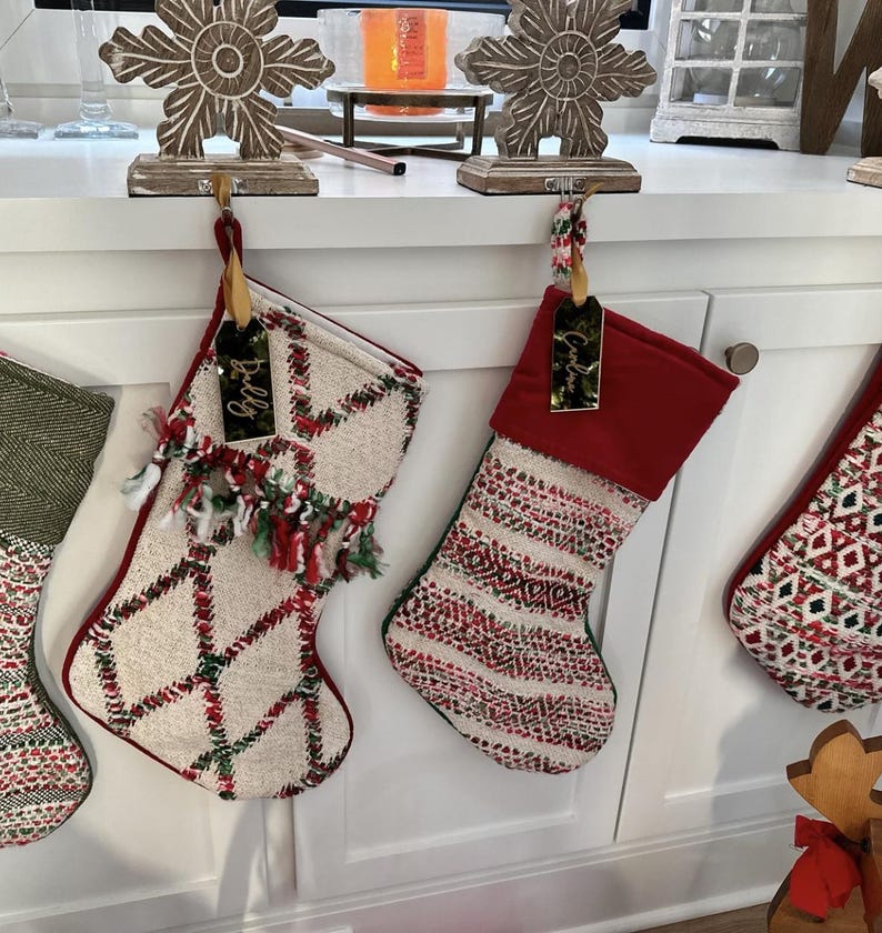 Op de afbeelding: Twee kerstsokken hangend aan een wit oppervlak. De linker sok is cr&egrave;mekleurig met een rood en groen ruitpatroon en franjes. De rechter sok heeft een effen rode boord en een cr&egrave;mekleurig lichaam met rode en groene geweven geometrische strepen.
