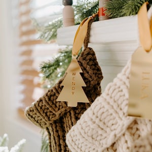 May include: Close-up of two Christmas stockings with gold name tags. One stocking is brown, the other is cream-colored. The gold tag on the brown stocking reads "WENDY" in capital letters. The tags are shaped like Christmas trees.