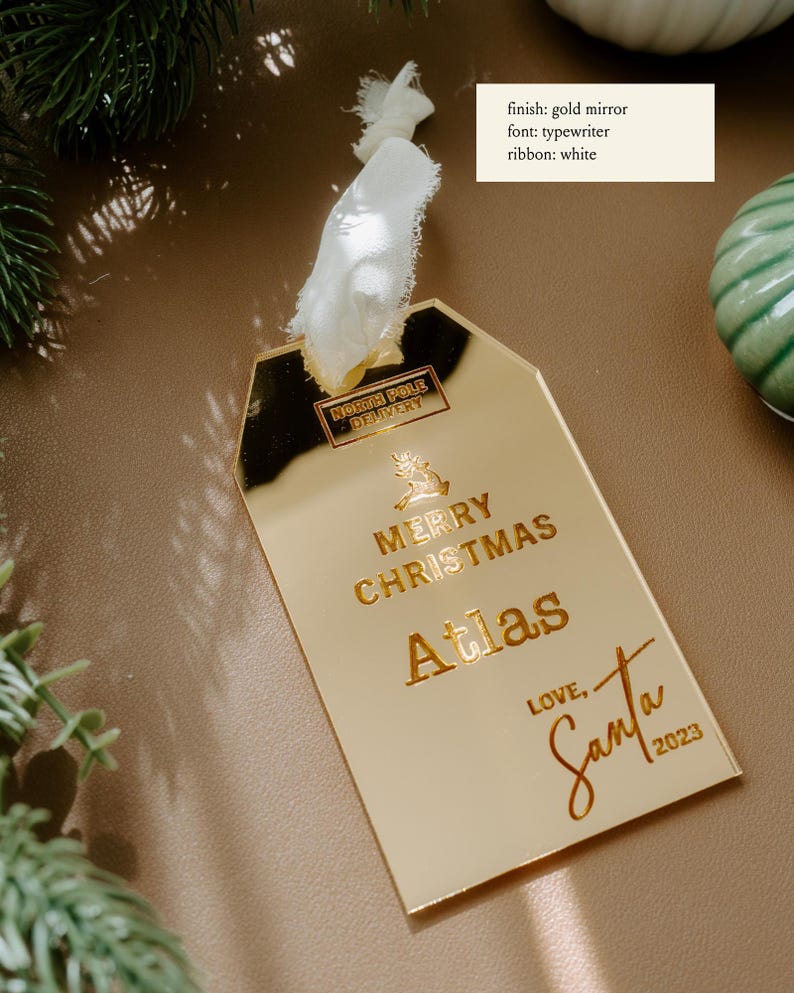 May include: Gold mirror Christmas ornament with the text "NORTH POLE DELIVERY", "MERRY CHRISTMAS", "Atlas", "LOVE, Santa", and the year "2023".