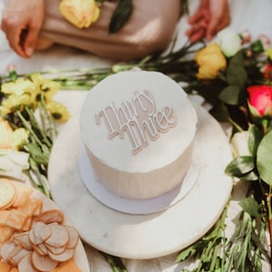 Pode incluir: Um bolo branco com as palavras "Thirty Three" escritas em glacê rosa no topo. O bolo está em um prato branco e rodeado de flores.
