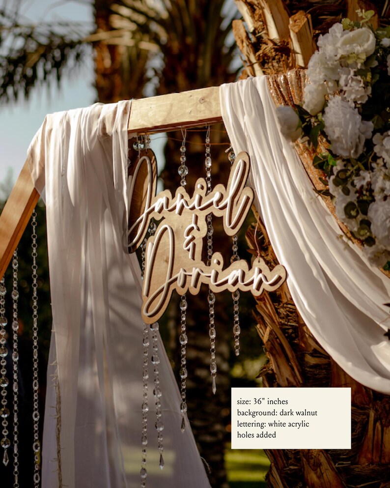 Puede incluir: Un letrero de boda de madera con los nombres "Daniel & Dorian" en escritura cursiva. El letrero tiene un tama&ntilde;o de 91,44 cm y un fondo de nogal oscuro con letras de acr&iacute;lico blanco. Se han a&ntilde;adido agujeros al letrero.