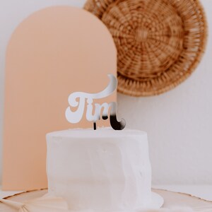 May include: A white cake with a silver cake topper that says "Tim" in a retro font. The cake is on a wooden cake stand with a woven basket and a peach-colored arch in the background.