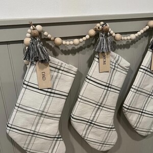 Op de afbeelding: Een slinger van natuurlijke houten kralen met hangende kerstsokken van off-white stof met een zwart en grijs ruitpatroon. Elke sok is voorzien van een grijze kwast en een houten label met de tekst DAD of MOM.