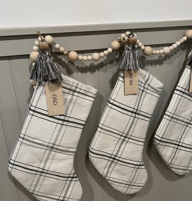 Op de afbeelding: Een slinger van natuurlijke houten kralen met hangende kerstsokken van off-white stof met een zwart en grijs ruitpatroon. Elke sok is voorzien van een grijze kwast en een houten label met de tekst DAD of MOM.