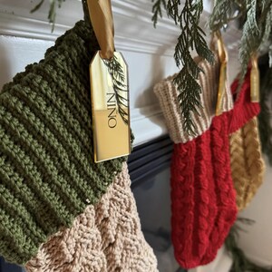 Op de afbeelding: Een set van drie gebreide kerstsokken die op een rij hangen. De eerste is olijfgroen en cr&egrave;me met een gouden spiegel-acryl label met de tekst NINO. De tweede is rood met een cr&egrave;me boord en de derde is goudkleurig. Alle hebben een kabelpatroon.