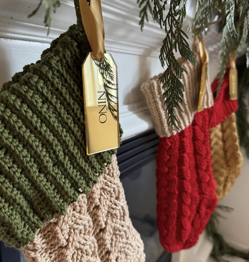 Op de afbeelding: Een set van drie gebreide kerstsokken die op een rij hangen. De eerste is olijfgroen en cr&egrave;me met een gouden spiegel-acryl label met de tekst NINO. De tweede is rood met een cr&egrave;me boord en de derde is goudkleurig. Alle hebben een kabelpatroon.