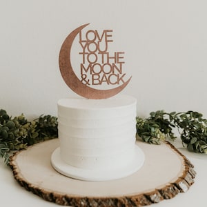 May include: A white cake with a wooden moon and back topper that says "Love you to the moon & back". The cake is on a wooden cake stand.