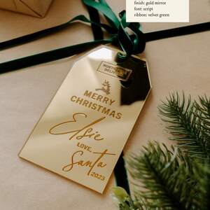 May include: Gold mirror gift tag with the text "Merry Christmas Elsie Love, Santa 2023" in a script font. The tag is tied with a green velvet ribbon.
