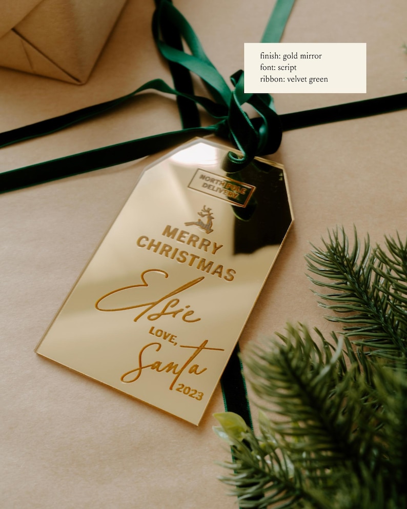 May include: Gold mirror gift tag with the text "Merry Christmas Elsie Love, Santa 2023" in a script font. The tag is tied with a green velvet ribbon.