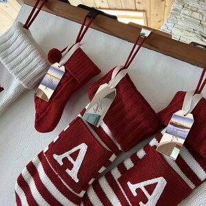 Op de afbeelding: Een verzameling gebreide kerstsokken hangend aan een houten schoorsteenmantel. De sokken hebben rood-witte strepen of effen rode breipatronen met witte letter A applicaties. Elke sok is voorzien van een reflecterend zilverkleurig cadeaulabel.