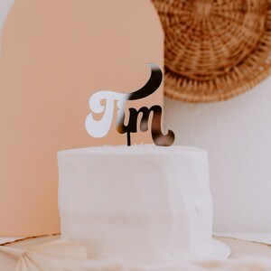 May include: A white cake with a black and white "Jim" cake topper. The cake is on a wooden cake stand.