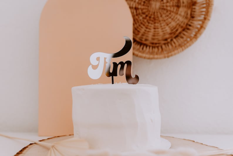 May include: A white cake with a black and white "Jim" cake topper. The cake is on a wooden cake stand.