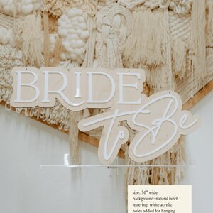 Puede incluir: Un letrero de madera con las palabras "Bride to Be" en letras acr&iacute;licas blancas sobre un fondo de abedul natural. El letrero tiene 91,44 cm de ancho y tiene agujeros para colgar.