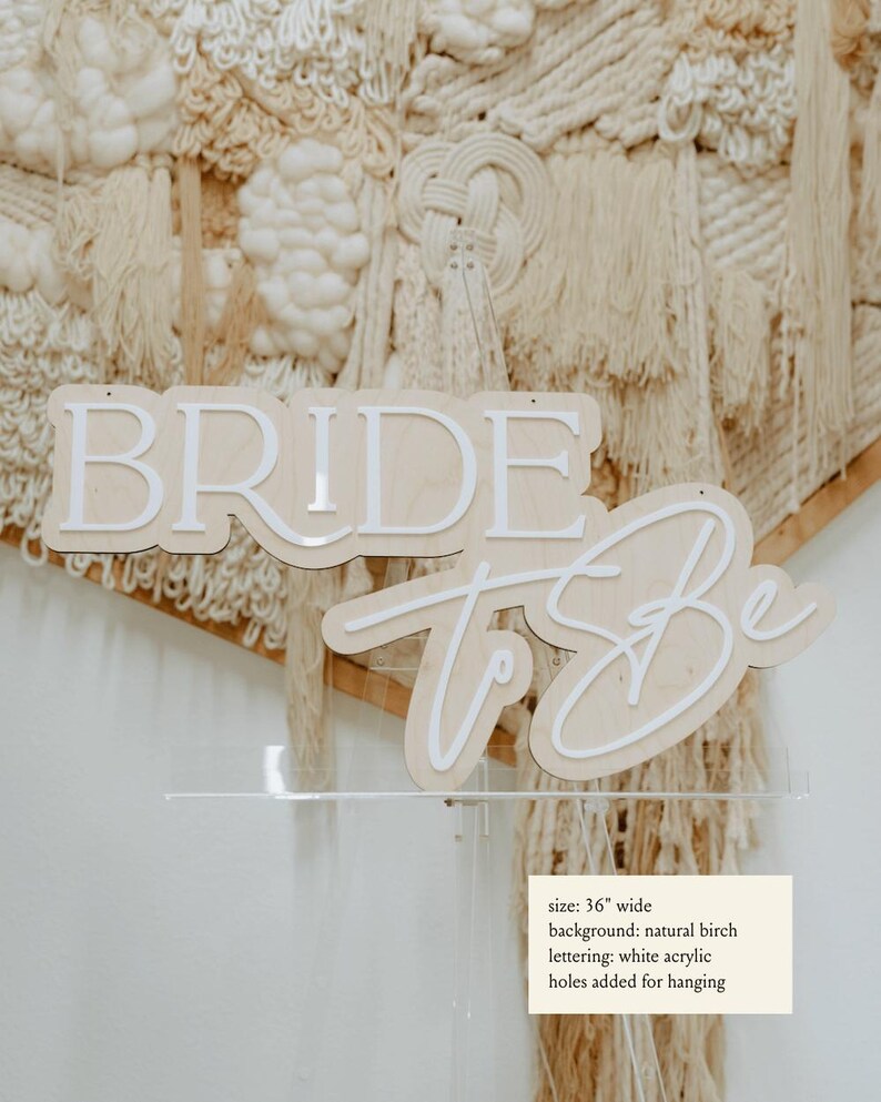 Puede incluir: Un letrero de madera con las palabras "Bride to Be" en letras acr&iacute;licas blancas sobre un fondo de abedul natural. El letrero tiene 91,44 cm de ancho y tiene agujeros para colgar.