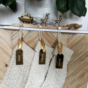 Op de afbeelding: Drie cr&egrave;mekleurige gebreide kerstsokken hangen aan een houten houder met een goudkleurige metalen arrenslee en twee rendieren. Elke sok is versierd met een gouden lint en een rechthoekig gouden spiegellabel met de namen John, Melina en Beans.