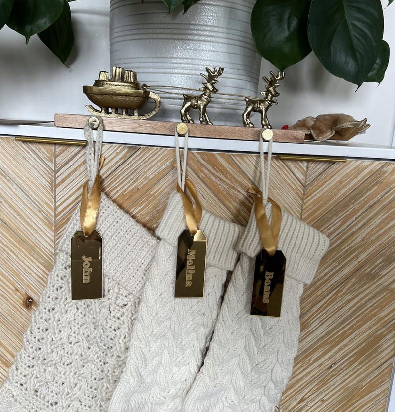 Op de afbeelding: Drie cr&egrave;mekleurige gebreide kerstsokken hangen aan een houten houder met een goudkleurige metalen arrenslee en twee rendieren. Elke sok is versierd met een gouden lint en een rechthoekig gouden spiegellabel met de namen John, Melina en Beans.