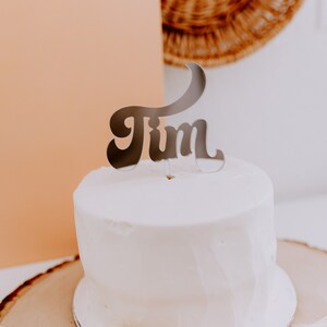 May include: A white cake with a silver "Jim" cake topper on top. The cake is on a wooden cake stand and there is a white fan behind it.