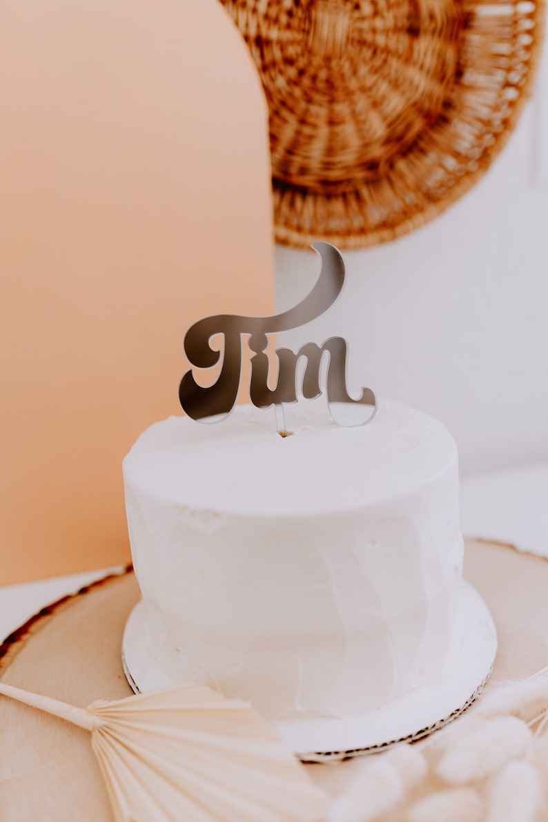 May include: A white cake with a silver "Jim" cake topper on top. The cake is on a wooden cake stand and there is a white fan behind it.