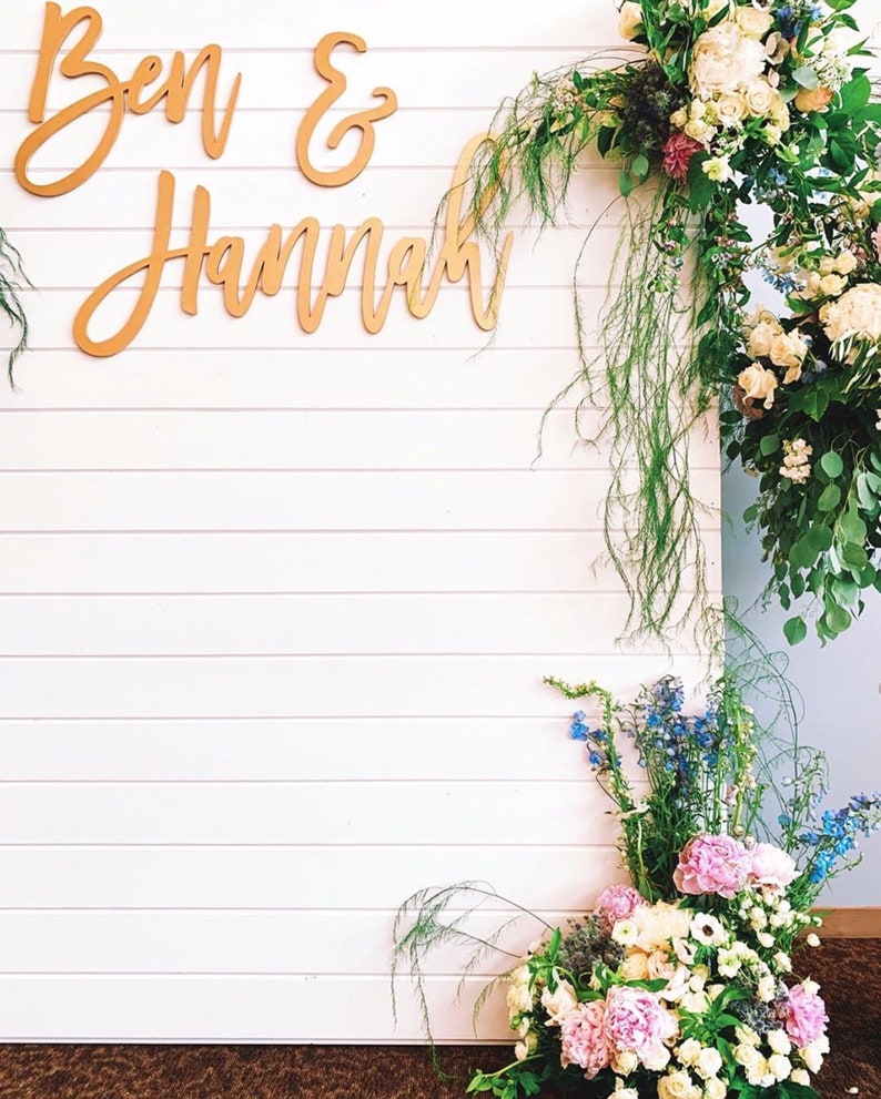 May include: White wooden backdrop with a gold sign that reads "Ben & Hannah" and a floral arrangement with pink, white, and blue flowers.