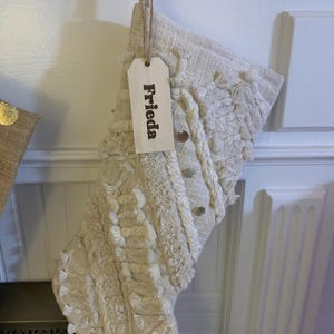 Op de afbeelding: Een cr&egrave;mekleurige, gestructureerde kerstkous met geweven patronen, franjes en kleine metalen, ronde accenten. Een lichtgekleurd, rechthoekig houten cadeaulabel is aan de bovenkant bevestigd met touw en toont de naam Frieda in zwarte letters.