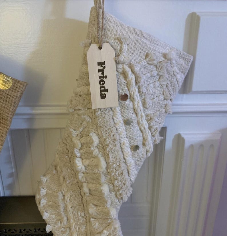 Op de afbeelding: Een cr&egrave;mekleurige, gestructureerde kerstkous met geweven patronen, franjes en kleine metalen, ronde accenten. Een lichtgekleurd, rechthoekig houten cadeaulabel is aan de bovenkant bevestigd met touw en toont de naam Frieda in zwarte letters.