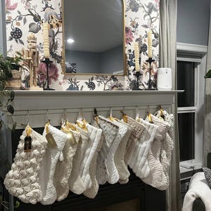 Op de afbeelding: Een rij van twaalf witte en cr&egrave;mekleurige kerstsokken hangend aan een witte schoorsteenmantel. De sokken hebben verschillende texturen, waaronder kabelbreiwerk, imitatiebont en pomponpatronen, elk voorzien van een klein goudkleurig naamplaatje.