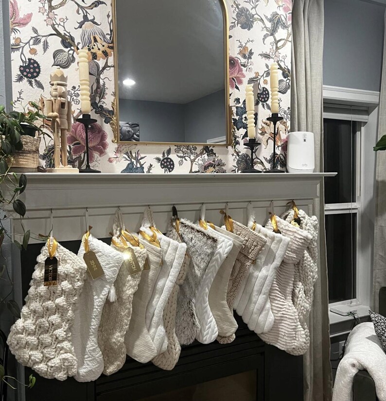 Op de afbeelding: Een rij van twaalf witte en cr&egrave;mekleurige kerstsokken hangend aan een witte schoorsteenmantel. De sokken hebben verschillende texturen, waaronder kabelbreiwerk, imitatiebont en pomponpatronen, elk voorzien van een klein goudkleurig naamplaatje.
