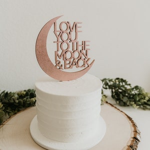 May include: A white cake with a wooden moon and back topper that says "Love you to the moon & back". The cake is on a wooden cake stand.