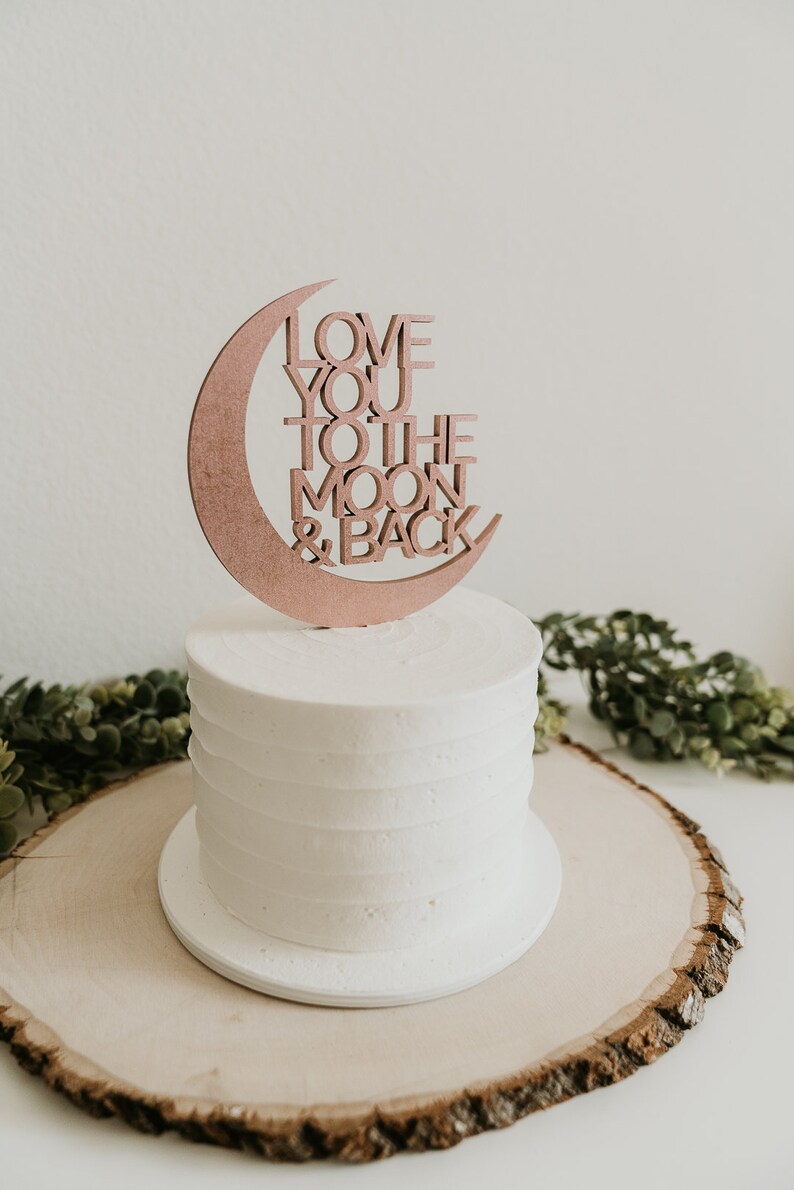 May include: A white cake with a wooden moon and back topper that says "Love you to the moon & back". The cake is on a wooden cake stand.