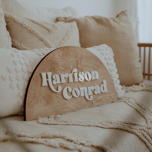 Puede incluir: Un letrero de madera en forma de semic&iacute;rculo con los nombres "Harrison Conrad" en letras blancas en relieve. El letrero est&aacute; sobre una cama con almohadas blancas y beige.