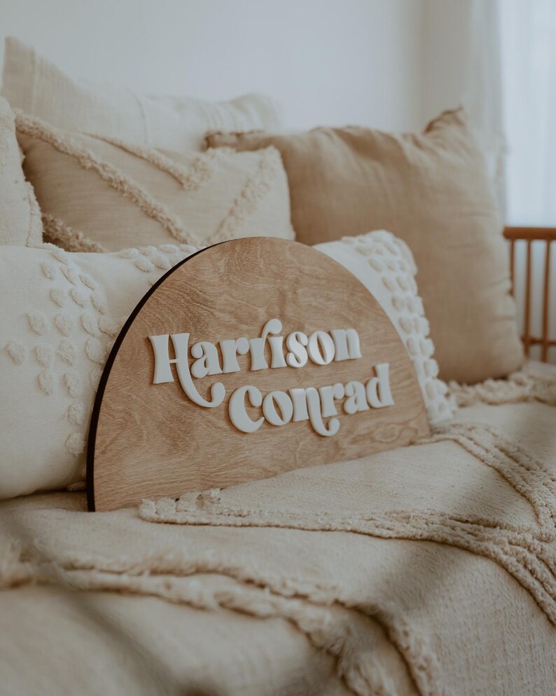 Puede incluir: Un letrero de madera en forma de semic&iacute;rculo con los nombres "Harrison Conrad" en letras blancas en relieve. El letrero est&aacute; sobre una cama con almohadas blancas y beige.