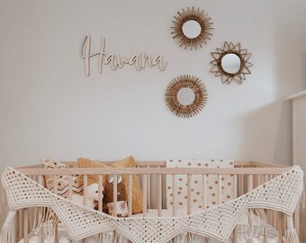 May include: A white crib with a crocheted garland and a wooden name sign that says "Havana". There are three woven star-shaped mirrors on the wall behind the crib.