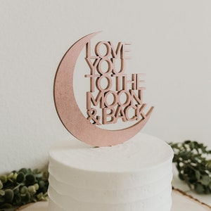 May include: A white cake with a wooden cake topper in the shape of a crescent moon with the words "Love You To The Moon & Back" written on it.